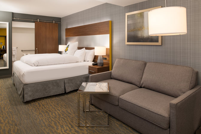 Modern hotel suite interior with a large bed, gray upholstered sofa, bedside lamps, sliding wooden barn door, framed artwork, and a small glass-top side table in neutral gray and wood decor.