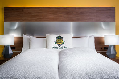 Bright modern hotel bedroom with a made king bed in white quilts, decorative pillow featuring a pineapple and the text “Sleep with Me,” wood-and-metal headboard, yellow accent wall and matching bedside lamps.