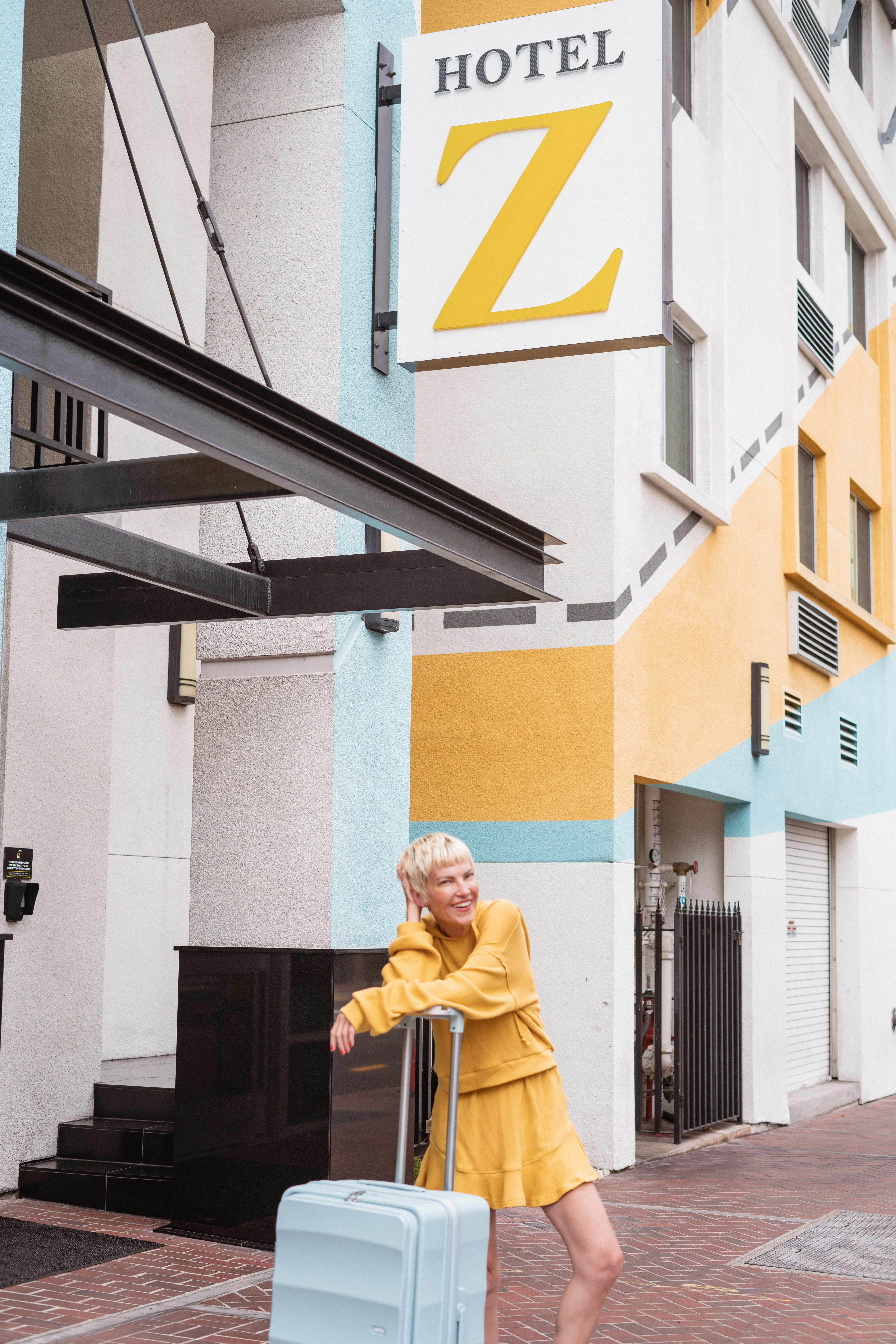 Smiling traveler in a yellow outfit leaning on a light-blue suitcase outside a colorful downtown boutique hotel with geometric yellow and teal facade.