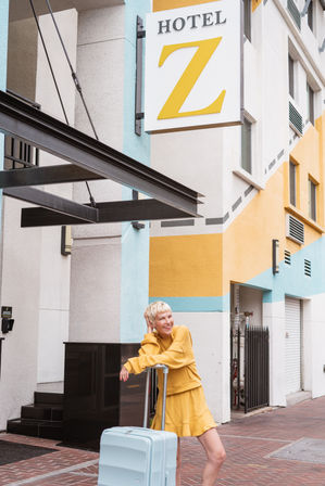 Smiling traveler in a yellow outfit leaning on a light-blue suitcase outside a colorful downtown boutique hotel with geometric yellow and teal facade.