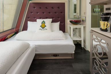 Cozy boutique micro-suite with tufted burgundy headboard, crisp white linens and pineapple throw pillow reading 'Sleep with Me', angled window, glass vessel sink vanity and counter-top coffee maker.