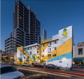 Dusk downtown street with a colorful yellow-and-blue low-rise hotel featuring a pineapple logo, parked cars and light trails, and a tall modern apartment high-rise towering behind.