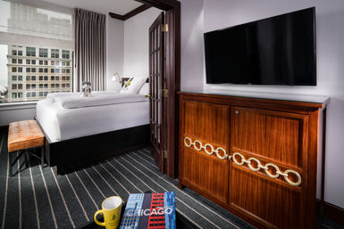 Cozy downtown Chicago hotel room with city-view window, white bed topped by a small stuffed animal, tufted leather bench, striped carpet, wooden cabinet with circular brass accents and wall-mounted TV, and a Chicago guidebook and yellow mug on the table.