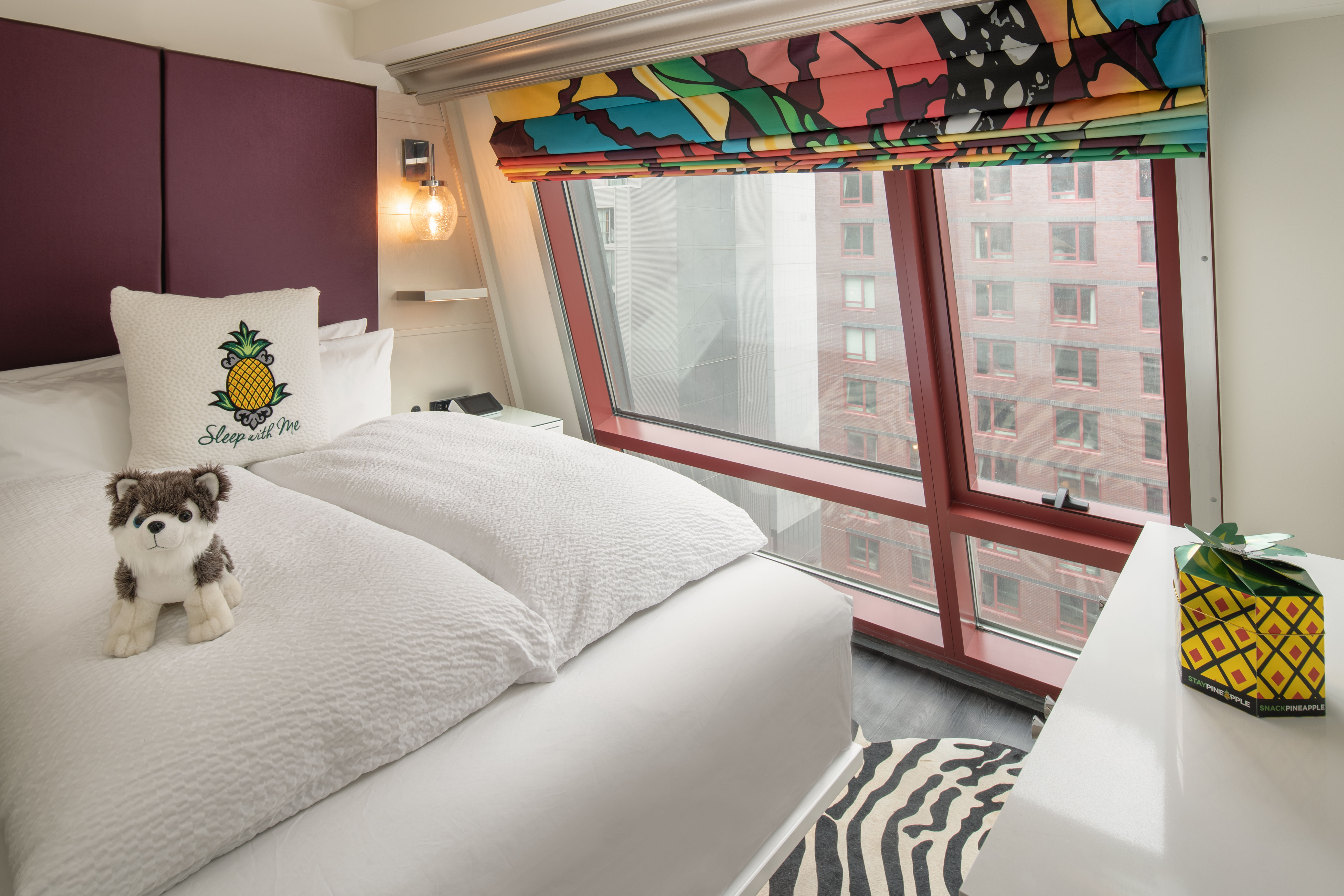 Cozy high-rise hotel bedroom with white bedding, plush husky on the bed, pineapple-printed pillow, colorful roman shade and angled city-view windows.