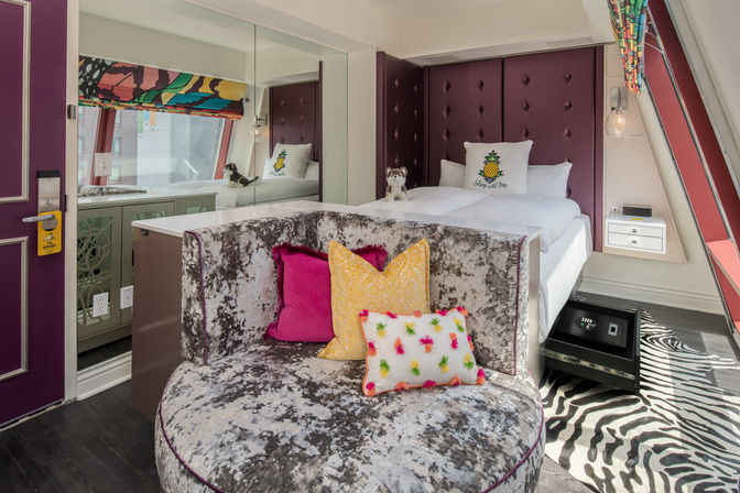 Plush boutique hotel room interior with crushed-velvet round sofa, colorful pillows including a pineapple motif, tufted purple headboard, zebra-print rug and angled city-view window.