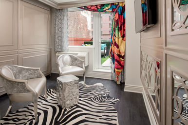 Stylish urban sitting area with two silver upholstered chairs, a metallic cube side table on a zebra-print rug, and a bold multicolor curtain framing a glass door to a small balcony and brick city view.