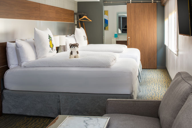 Bright modern hotel room with two neatly made beds in white linens, pineapple-embroidered pillows, a cute plush husky on the near bed, sliding wooden barn door to the bathroom, gray armchair, wall-mounted TV and natural light from a window.