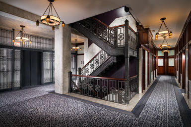 Historic hotel interior with ornate wrought-iron staircase, vintage gated elevators, marble columns, patterned carpeted corridors, and warm hanging chandeliers.