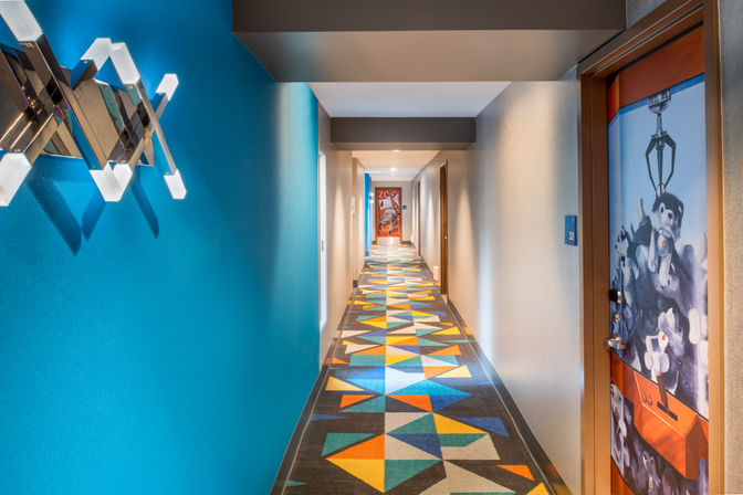 Vibrant hotel corridor with teal accent wall, contemporary wall lights, colorful geometric carpet, and a guest-room door featuring a playful claw-machine stuffed-animal mural.