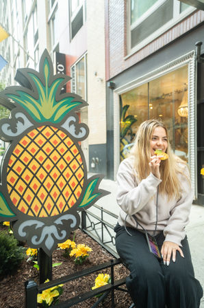 Woman sitting on a downtown sidewalk beside a large decorative pineapple sign, smiling while eating a pineapple slice outside an urban storefront with yellow flowers