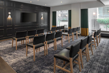 Sleek urban meeting room with rows of black upholstered chairs facing a wall-mounted TV on a dark paneled accent wall, large windows with green-and-gold curtains overlooking a city street, and textured gray patterned carpet.