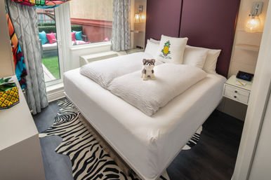 Cozy boutique hotel bedroom with neatly made white bed, small stuffed husky on the duvet, pineapple decorative pillow, zebra-striped rug and sliding glass door leading to a balcony with colorful cushions.