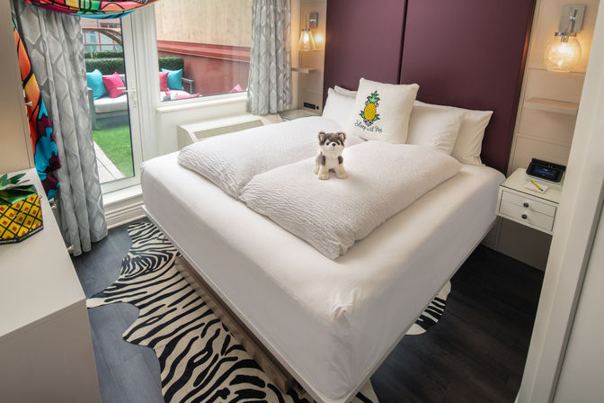 Cozy boutique hotel bedroom with neatly made white bed, small stuffed husky on the duvet, pineapple decorative pillow, zebra-striped rug and sliding glass door leading to a balcony with colorful cushions.