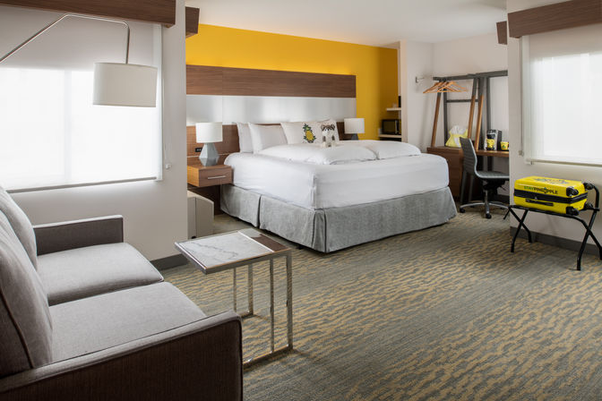 Spacious modern hotel room with king bed and white linens, cheerful yellow accent wall, bedside lamps, sofa with marble-top side table, work desk and open wardrobe, and a bright yellow suitcase on a luggage rack.