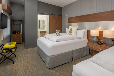 Cozy modern hotel room with two white beds, wood headboard, bedside lamps, pineapple accent pillow, plush husky on the bed, yellow pineapple suitcase on a luggage rack, and a sliding barn-door to the bathroom.