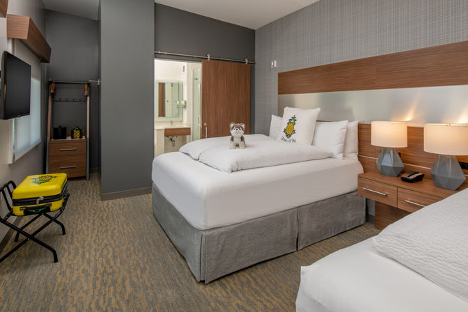 Cozy modern hotel room with two white beds, wood headboard, bedside lamps, pineapple accent pillow, plush husky on the bed, yellow pineapple suitcase on a luggage rack, and a sliding barn-door to the bathroom.