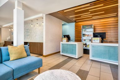 Bright modern hotel lobby with blue upholstered sofa and yellow pillow, marble coffee table, twin reception desks with glass-front panels, wood-slat accent ceiling and a stocked refrigerated snack and beverage display