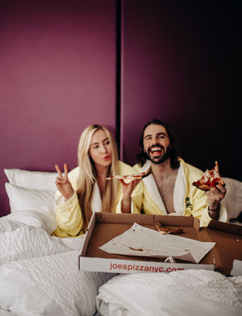 Couple in matching yellow robes laughing and eating pizza in bed during a cozy hotel staycation against a purple accent wall.