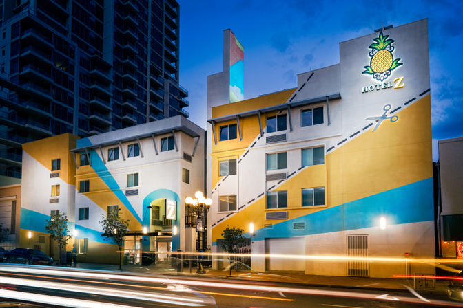 Colorful downtown boutique hotel at dusk with geometric yellow-and-blue facade, pineapple mural above entrance, streetlamp and streaking car light trails