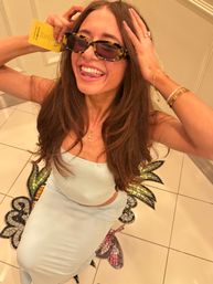 Playful indoor selfie of a smiling woman in a light blue two-piece dress and tortoiseshell sunglasses, sticking out her tongue while holding a yellow card, sitting on white tiled floor with a colorful mosaic pineapple motif and wearing gold jewelry.