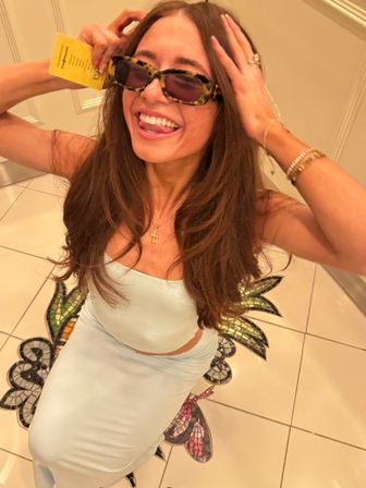 Playful indoor selfie of a smiling woman in a light blue two-piece dress and tortoiseshell sunglasses, sticking out her tongue while holding a yellow card, sitting on white tiled floor with a colorful mosaic pineapple motif and wearing gold jewelry.
