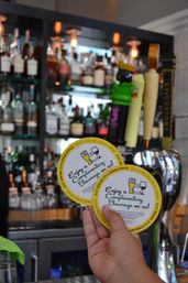 Hand holding two round coasters that read Enjoy a Complimentary Beverage on us! in front of a lively bar scene with draft taps and bottles on the back bar