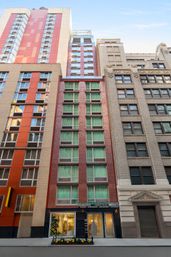 Tall, narrow red-brick mid-rise with rows of green-curtained windows and a glass storefront entrance on a downtown city street, flanked by orange-paneled and stone high-rises.