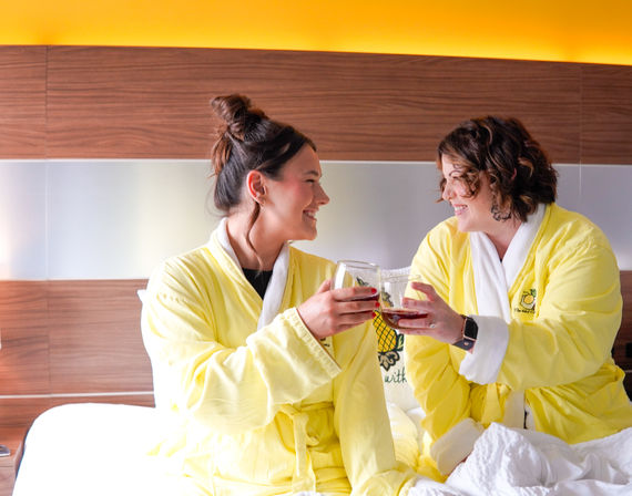 Two friends in bright yellow robes clinking wine glasses on a hotel bed with warm wood headboard, enjoying a cozy staycation.