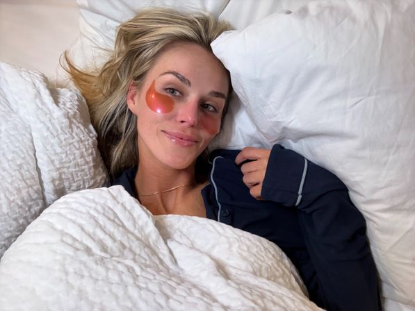 Smiling woman relaxing in a white bed with rose-gold hydrogel under-eye patches and a navy pajama top in a cozy skincare self-care scene