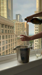 Hands pouring white wine into a stemless glass at an urban high-rise window, metal ice bucket on the sill and city skyline beyond