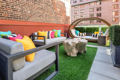 Cheerful urban rooftop patio with gray modern sofas and brightly colored throw pillows, a circular wicker daybed and live-edge wood coffee table on green turf beside a brick apartment building.