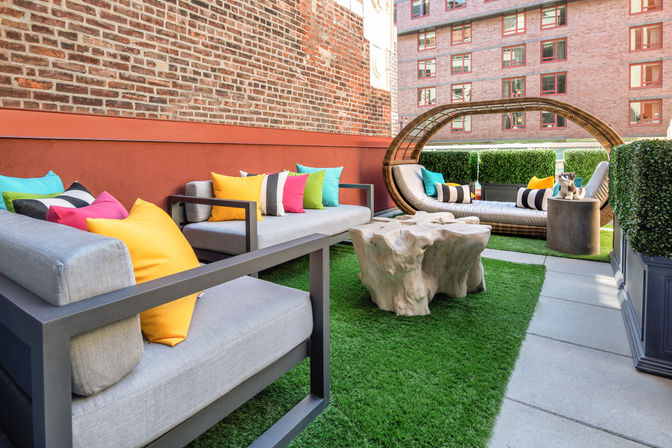 Cheerful urban rooftop patio with gray modern sofas and brightly colored throw pillows, a circular wicker daybed and live-edge wood coffee table on green turf beside a brick apartment building.