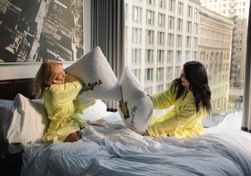 Two friends in yellow robes having a playful pillow fight on a rumpled hotel bed by a large window with downtown high‑rise buildings and white pillows featuring pineapple graphics.