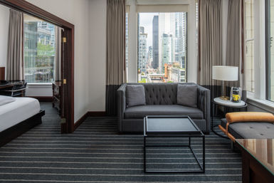 Modern downtown hotel suite living area with gray tufted sofa, striped carpet, black metal coffee table and adjacent bed, framed by a large window showcasing an urban high-rise skyline and city street below