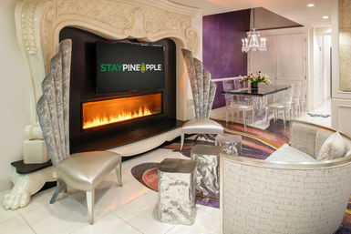 Glamorous boutique hotel lounge with ornate carved mantel and built-in linear fireplace beneath a wall-mounted TV, silver high-backed chairs, hammered-metal stools, curved upholstered sofa, glass dining table with clear chairs and crystal chandelier, purple accent wall and colorful patterned rug.