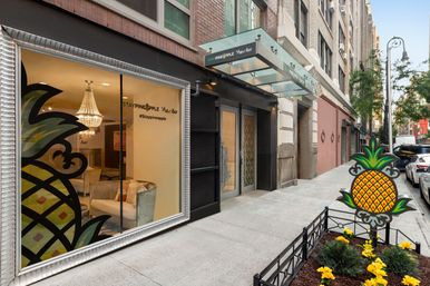 Street-level boutique hotel entrance on a downtown city sidewalk with glass canopy, large window revealing chandelier-lit lobby seating, decorative pineapple sign in a planted bed and parked cars along the street