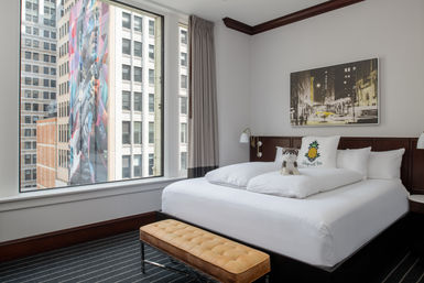 Bright downtown hotel bedroom with king bed, pineapple throw pillow and plush toy dog, tufted bench at foot of bed, large window with city view of high-rise buildings and a colorful street-art mural
