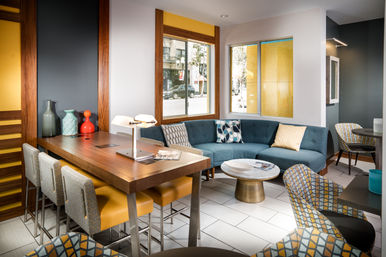 Bright contemporary urban lounge with curved teal sofa, patterned accent chairs, wooden high-top table with yellow stools and decorative vases, large windows looking onto a city street
