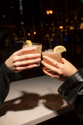 Two hands clinking lemon-garnished pink cocktails with ice and straws over a marble bar, warm city nightlife bokeh in the background — cheers to a night out.