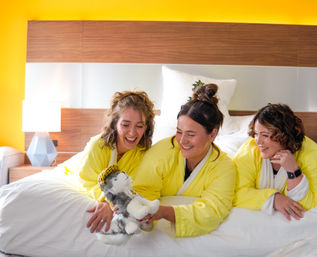 Three friends in bright yellow robes laughing on a hotel bed, playing with a plush husky toy against a modern wood headboard and warm yellow wall.