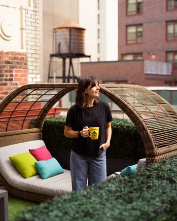 Smiling woman on an urban rooftop patio by a woven wicker daybed with bright pillows, holding a yellow pineapple mug with brick buildings and a rooftop water tower in the background.