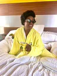 Smiling woman in a bright yellow pineapple-themed robe and cat-eye sunglasses sitting on a rumpled white bed in a modern hotel room, holding a pineapple-print mug