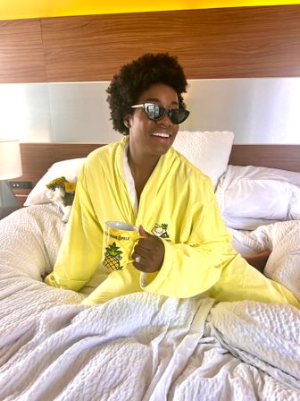 Smiling woman in a bright yellow pineapple-themed robe and cat-eye sunglasses sitting on a rumpled white bed in a modern hotel room, holding a pineapple-print mug