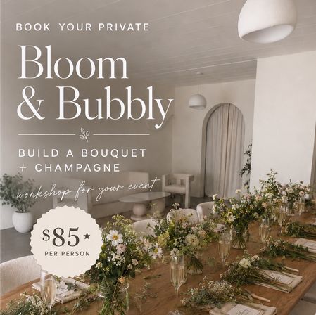 Private floral workshop setup in a bright minimalist studio: long wooden table with hand-tied wildflower bouquets, champagne flutes, place settings and neutral seating — "Bloom & Bubbly" event, $85 per person