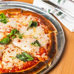 Close-up of a thin-crust Margherita pizza on a metal pan with melted mozzarella, vibrant basil leaves, tomato sauce and a slightly charred golden crust on a wooden table.
