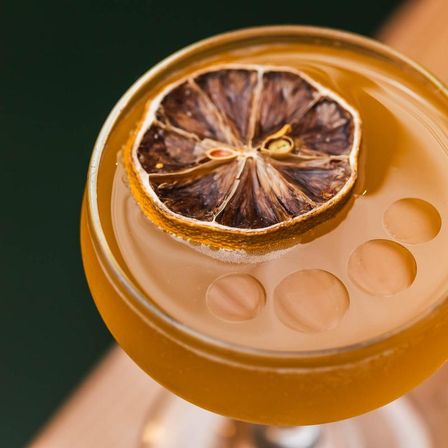Amber craft cocktail in a coupe glass garnished with a dried citrus wheel and three glossy foam bubbles
