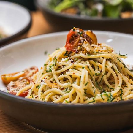 Close-up of Italian spaghetti aglio e olio with roasted cherry tomatoes, grated Parmesan, chopped parsley and red pepper flakes in a shallow bowl.