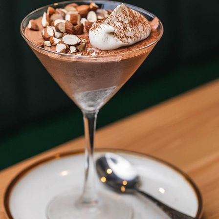 Decadent chocolate mousse in a martini glass topped with whipped cream and chopped hazelnuts, served on a white plate with a spoon on a wooden table