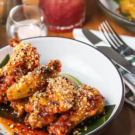 Mouthwatering sticky glazed chicken wings topped with crushed peanuts and black sesame seeds on a white plate with a lime wedge and cutlery on a restaurant table.