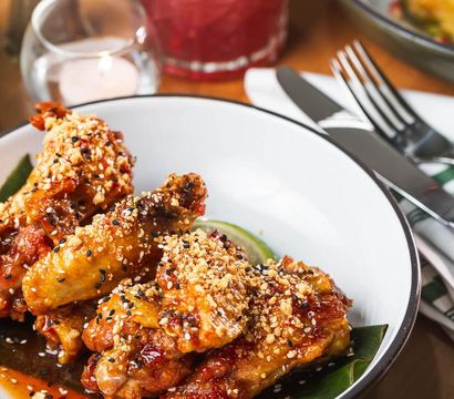Mouthwatering sticky glazed chicken wings topped with crushed peanuts and black sesame seeds on a white plate with a lime wedge and cutlery on a restaurant table.